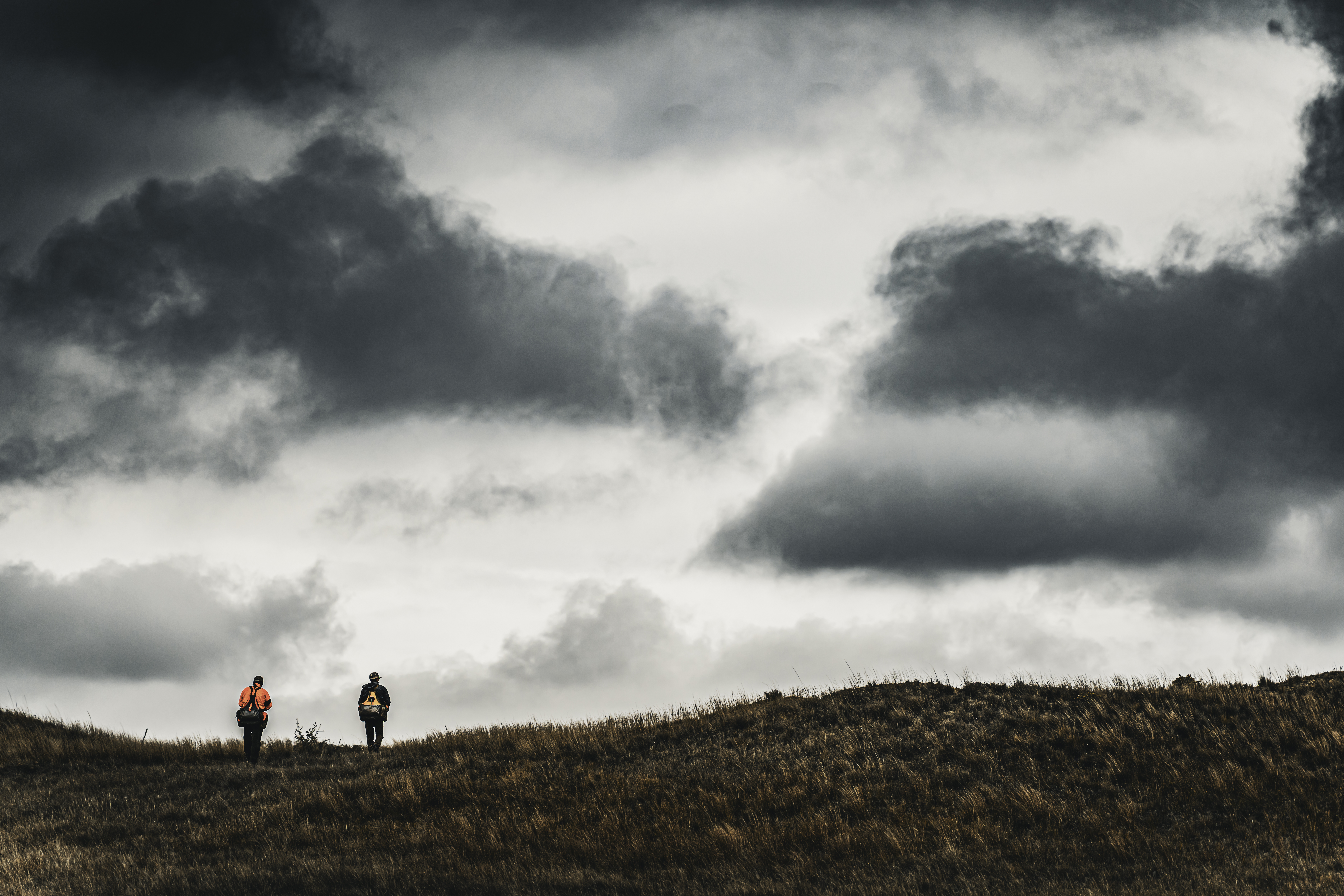 Two small figures walking across a windy ridge beneath a large cloud field.