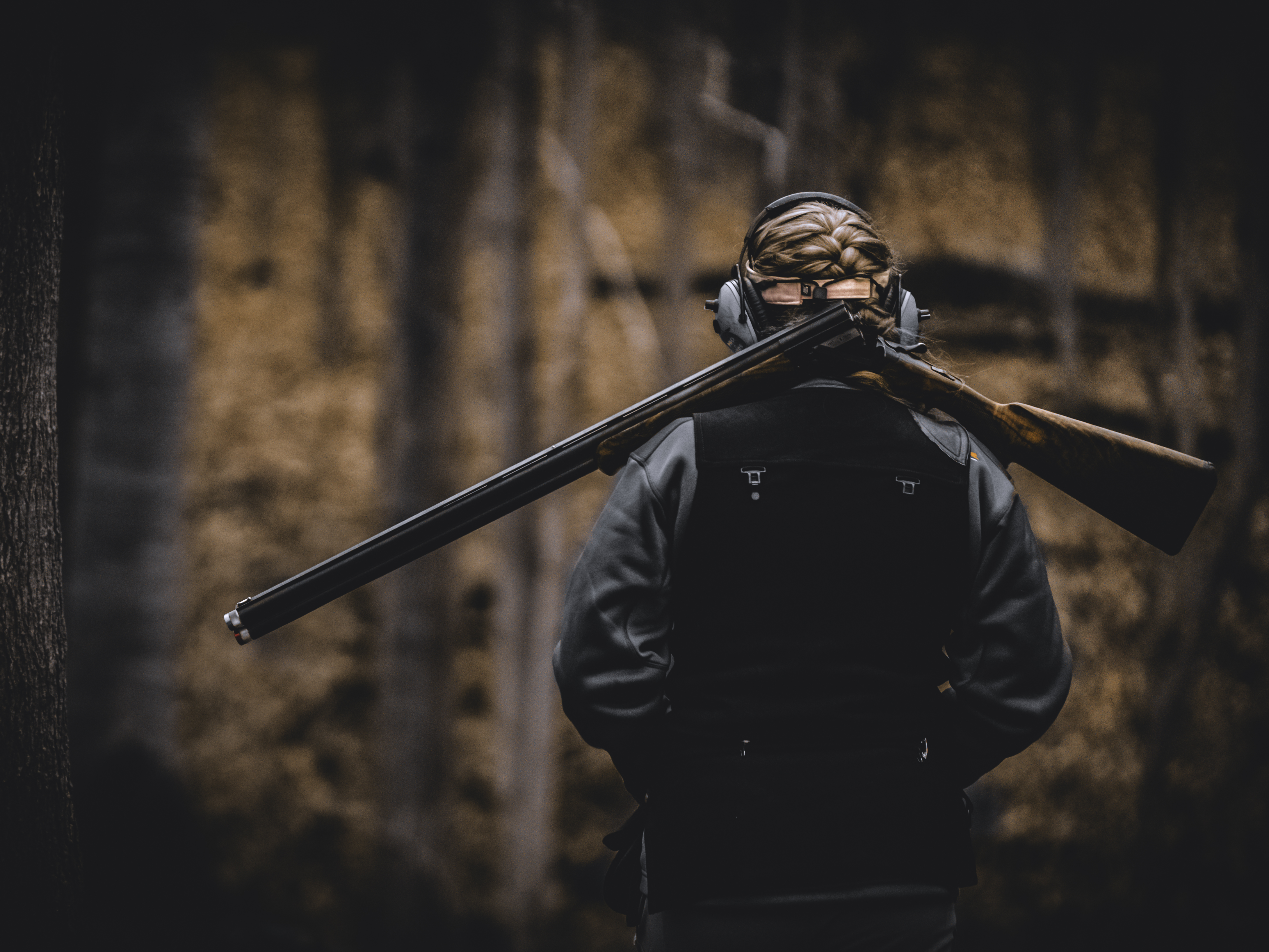 A person seen from behind in the woods with a shotgun resting across their shoulders.