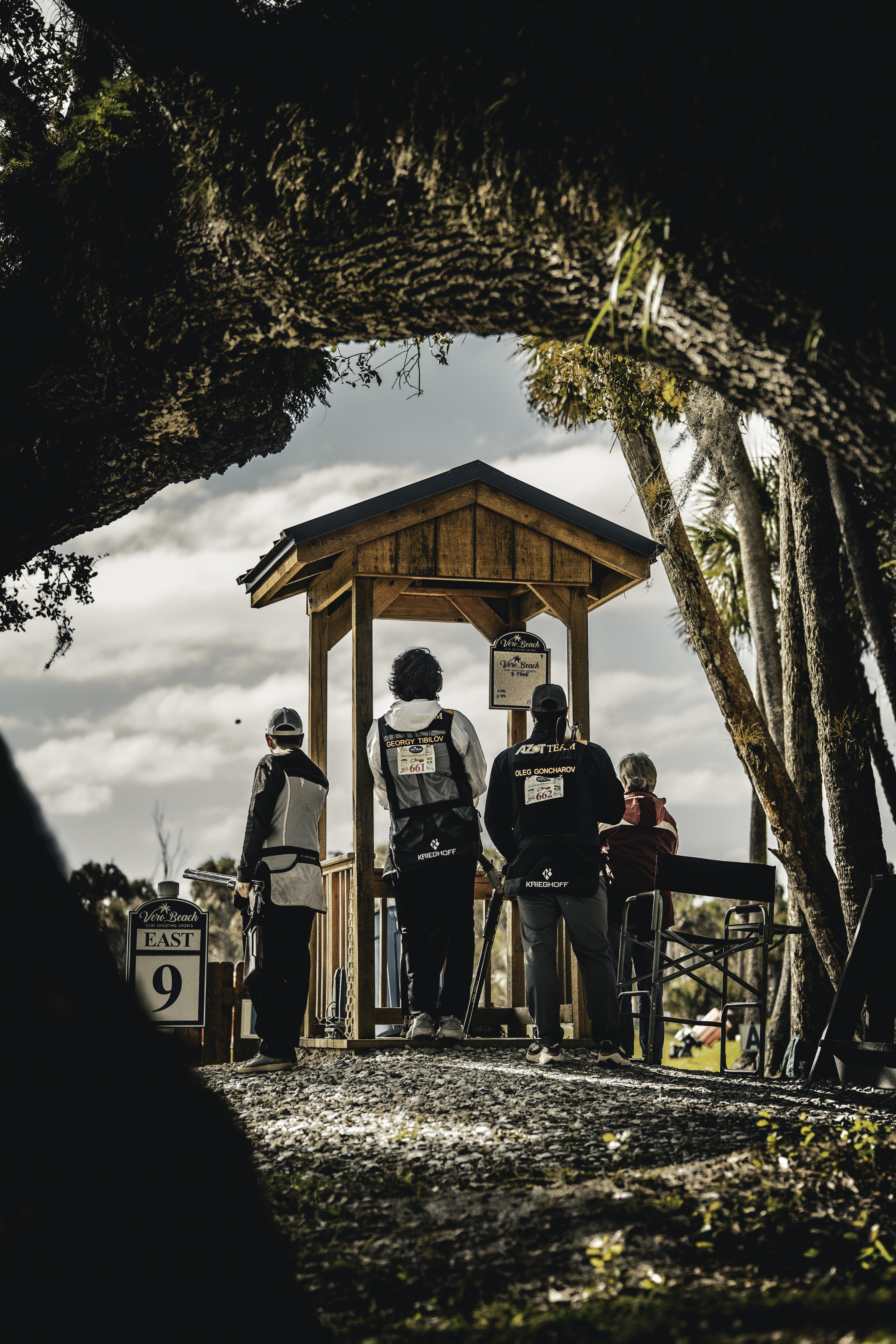 People standing near a shooting station beneath a dark tree canopy.